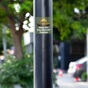 K&ouml;nnte beinhalten: Ein schwarzer Metallpfosten mit einem Aufkleber mit der Aufschrift "RESIST Only You Can Prevent Fascism". Der Aufkleber ist gr&uuml;n, gelb und braun. Der Hintergrund ist verschwommen und zeigt Gr&uuml;n und eine Stra&szlig;e.