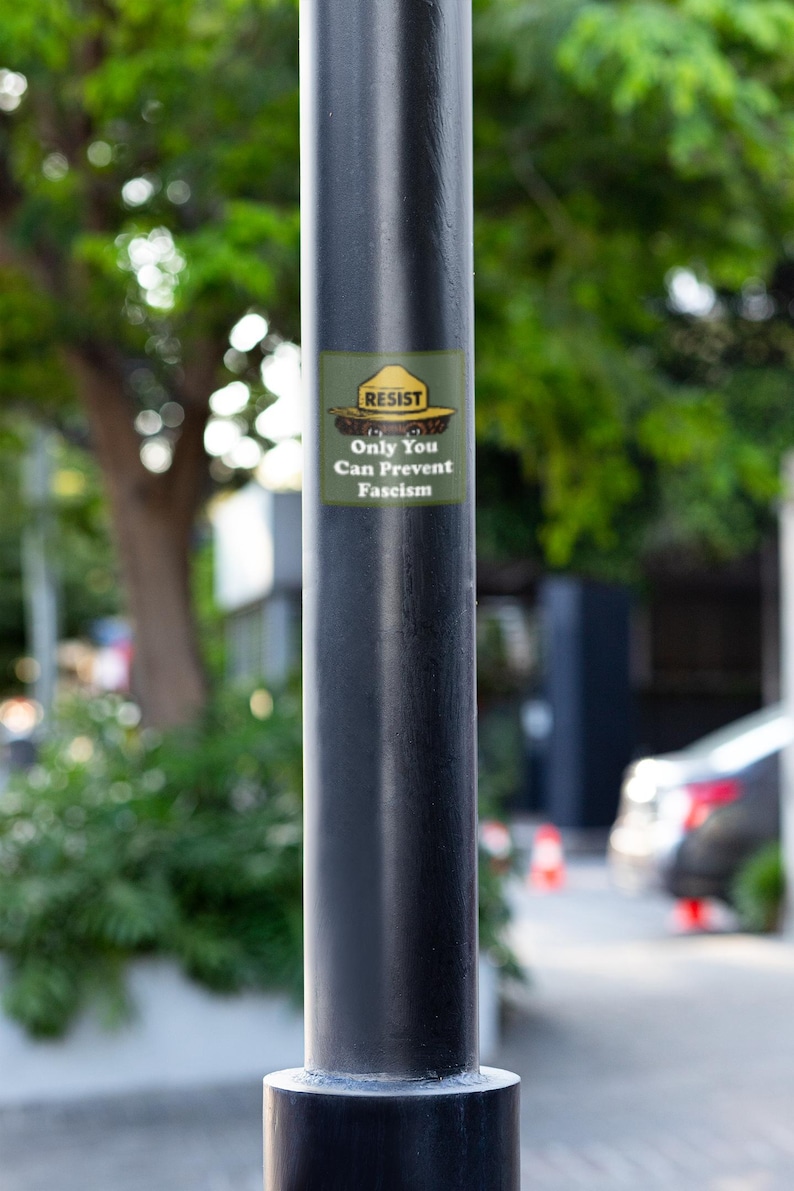 K&ouml;nnte beinhalten: Ein schwarzer Metallpfosten mit einem Aufkleber mit der Aufschrift "RESIST Only You Can Prevent Fascism". Der Aufkleber ist gr&uuml;n, gelb und braun. Der Hintergrund ist verschwommen und zeigt Gr&uuml;n und eine Stra&szlig;e.