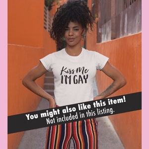 K&ouml;nnte beinhalten: Eine Person tr&auml;gt ein wei&szlig;es T-Shirt mit dem Aufdruck "Kiss Me I'M GAY". Die Person steht vor einer orangefarbenen Wand und tr&auml;gt eine gestreifte Hose.