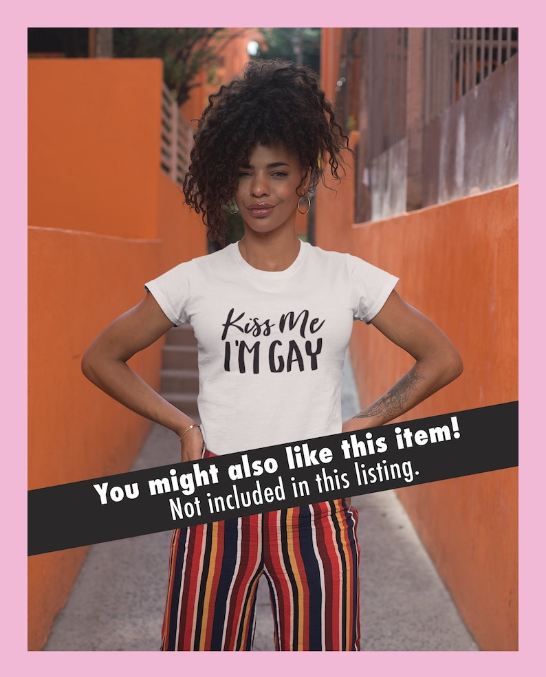 K&ouml;nnte beinhalten: Eine Person tr&auml;gt ein wei&szlig;es T-Shirt mit dem Aufdruck "Kiss Me I'M GAY". Die Person steht vor einer orangefarbenen Wand und tr&auml;gt eine gestreifte Hose.