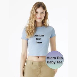 Op de afbeelding: Een lichtblauw Micro Rib Baby Tee met de tekst "custom text here" op de voorkant. De tee is een cropped stijl met korte mouwen. Het model draagt een kaki broek.