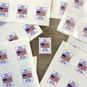 Pode incluir: Folhas de adesivos brancos com um design de selo postal. Cada selo apresenta uma bandeira americana vermelha, branca e azul, com o texto "PRO-AMERICA" acima e "ANTI-TRUMP" abaixo. Os adesivos estão espalhados em uma superfície de madeira.