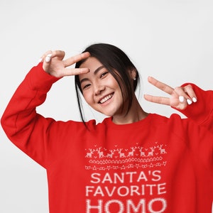 K&ouml;nnte beinhalten: Ein roter Weihnachtspullover mit einem wei&szlig;en Design, auf dem "Santas Lieblings-Homo" steht.
