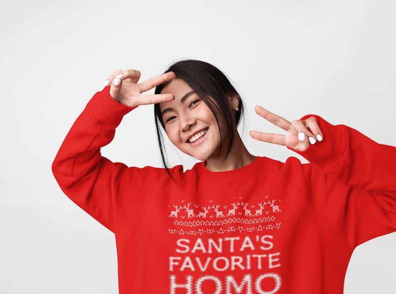 K&ouml;nnte beinhalten: Ein roter Weihnachtspullover mit einem wei&szlig;en Design, auf dem "Santas Lieblings-Homo" steht.