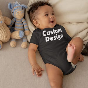May include: A baby wearing a black bodysuit with the text "Custom Design" in white letters. The baby is lying on a light brown couch with two crocheted stuffed animals.
