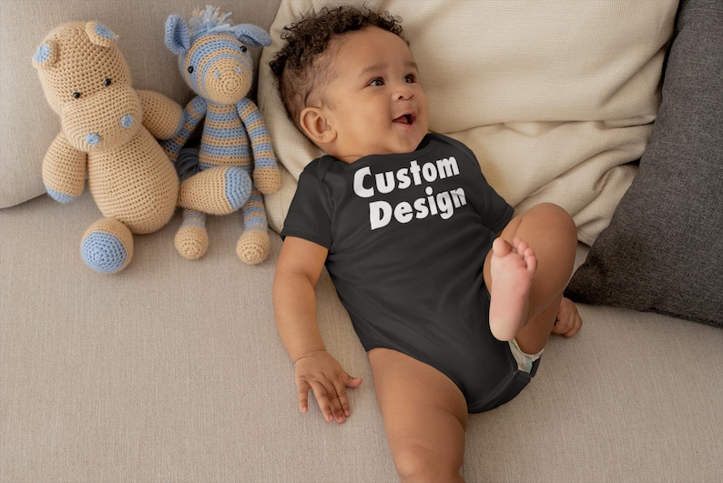May include: A baby wearing a black bodysuit with the text "Custom Design" in white letters. The baby is lying on a light brown couch with two crocheted stuffed animals.
