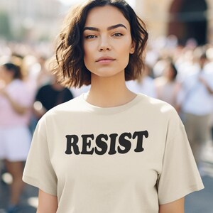 Pode incluir: Uma mulher vestindo uma camiseta bege claro com a palavra "RESIST" impressa em preto. Ela est&aacute; em p&eacute; na frente de uma multid&atilde;o de pessoas.