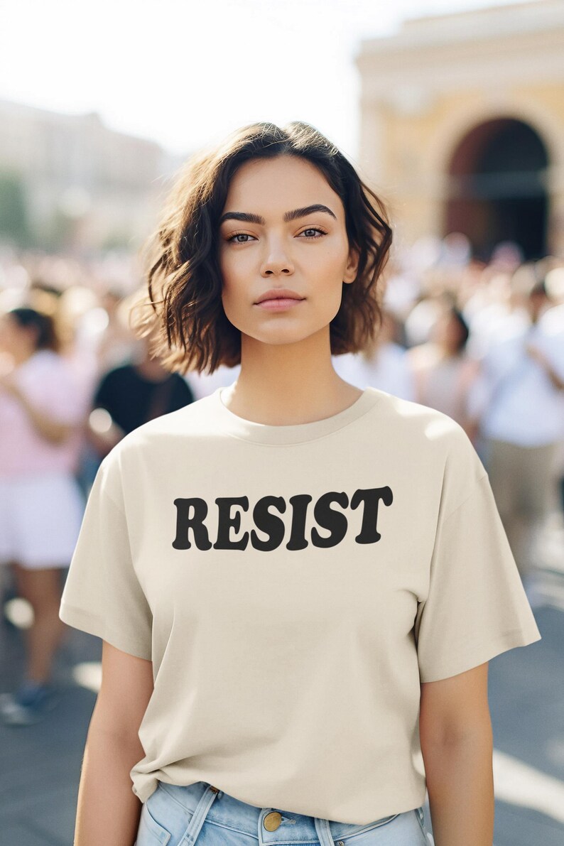 Pode incluir: Uma mulher vestindo uma camiseta bege claro com a palavra "RESIST" impressa em preto. Ela est&aacute; em p&eacute; na frente de uma multid&atilde;o de pessoas.
