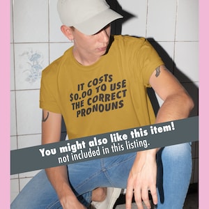 K&ouml;nnte beinhalten: Eine Person tr&auml;gt ein senfgelbes T-Shirt mit dem Aufdruck "IT COSTS 0,00 &euro; TO USE THE CORRECT PRONOUNS". Die Person tr&auml;gt eine hellgraue Baseballkappe und eine blaue Jeans.