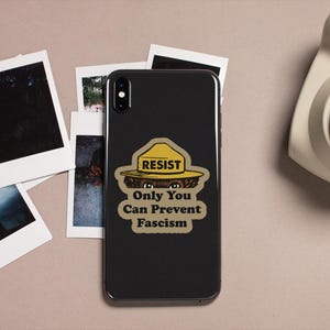 K&ouml;nnte beinhalten: Ein schwarzes Smartphone mit einem Aufkleber mit der Aufschrift "RESIST Only You Can Prevent Fascism" ist von Polaroid-Fotos und einer Vintage-Kamera umgeben. Der Aufkleber zeigt einen gelben Hut und einen B&auml;ren.