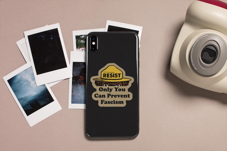 K&ouml;nnte beinhalten: Ein schwarzes Smartphone mit einem Aufkleber mit der Aufschrift "RESIST Only You Can Prevent Fascism" ist von Polaroid-Fotos und einer Vintage-Kamera umgeben. Der Aufkleber zeigt einen gelben Hut und einen B&auml;ren.