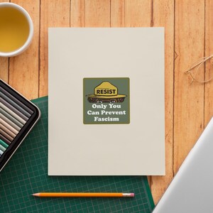 K&ouml;nnte beinhalten: Eine cremefarbene Karte mit einer gr&uuml;n-gelben Grafik mit der Aufschrift "RESIST Only You Can Prevent Fascism". Die Karte liegt auf einer Holzoberfl&auml;che mit einer Tasse Tee, einem Etui mit Buntstiften, einem Laptop, einem Bleistift und einer Brille.