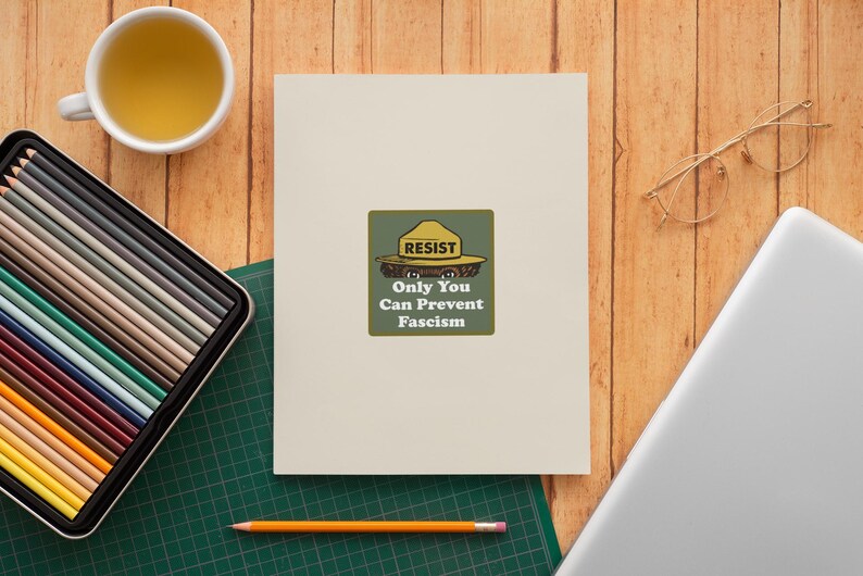 K&ouml;nnte beinhalten: Eine cremefarbene Karte mit einer gr&uuml;n-gelben Grafik mit der Aufschrift "RESIST Only You Can Prevent Fascism". Die Karte liegt auf einer Holzoberfl&auml;che mit einer Tasse Tee, einem Etui mit Buntstiften, einem Laptop, einem Bleistift und einer Brille.
