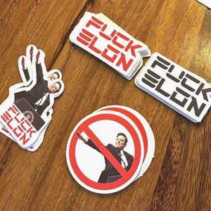 Pode incluir: Um monte de adesivos com o texto "FUCK ELON" em letras pretas e vermelhas sobre um fundo branco. Há também um adesivo com um círculo vermelho e uma linha vermelha diagonal que o atravessa, com uma imagem de Elon Musk dentro do círculo. Os adesivos estão sobre uma superfície de madeira.
