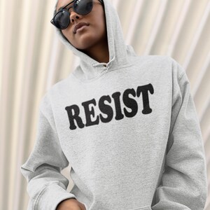 Pode incluir: Uma camisa de moletom cinza com capuz com a palavra "RESIST" impressa em letras pretas na frente.