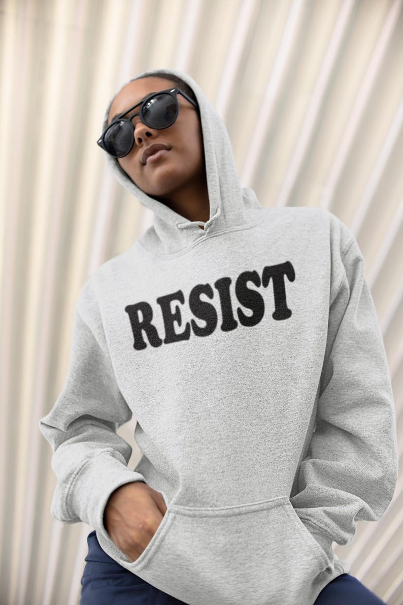 Pode incluir: Uma camisa de moletom cinza com capuz com a palavra "RESIST" impressa em letras pretas na frente.
