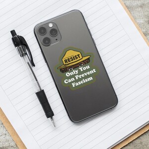 K&ouml;nnte beinhalten: Ein schwarzes Smartphone mit einem Aufkleber, der einen Cartoon-B&auml;ren mit gelbem Hut, das Wort "RESIST" und den Text "Only You Can Prevent Fascism" zeigt. Ein schwarzer Stift liegt auf einem linierten Notizblock.