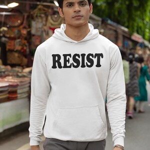 Pode incluir: Uma camisa de moletom branca com capuz com a palavra "RESIST" impressa em letras pretas e em negrito na frente. O modelo est&aacute; vestindo a camisa de moletom e cal&ccedil;as cinza.