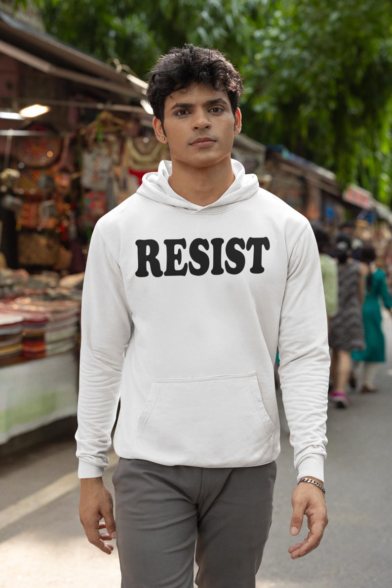 Pode incluir: Uma camisa de moletom branca com capuz com a palavra "RESIST" impressa em letras pretas e em negrito na frente. O modelo est&aacute; vestindo a camisa de moletom e cal&ccedil;as cinza.