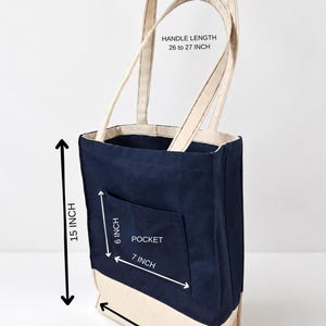 Puede incluir: Un bolso tote de lona azul marino y beige con un bolsillo frontal. Las dimensiones son 38,1 cm de alto, 27,9 cm de ancho y 10,2 cm de profundidad. Tiene asas de 66 a 68,6 cm. El texto dice Tailored by Faiza.