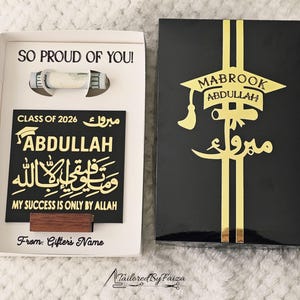 May include: A graduation gift box with a black and gold design. The box is open, revealing a USB drive and a black plaque with gold lettering that reads "ABDULLAH" and "CLASS OF 2026". The box lid has the words "MABROOK ABDULLAH" in gold.