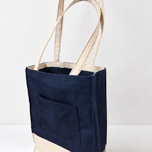 Puede incluir: Un bolso tote de lona en azul marino y crema con dos asas largas para el hombro, un bolsillo exterior rectangular en la parte delantera y una base s&oacute;lida de color crema. En la esquina inferior izquierda aparece el texto Tailored by Faiza Handcrafted Boutique.