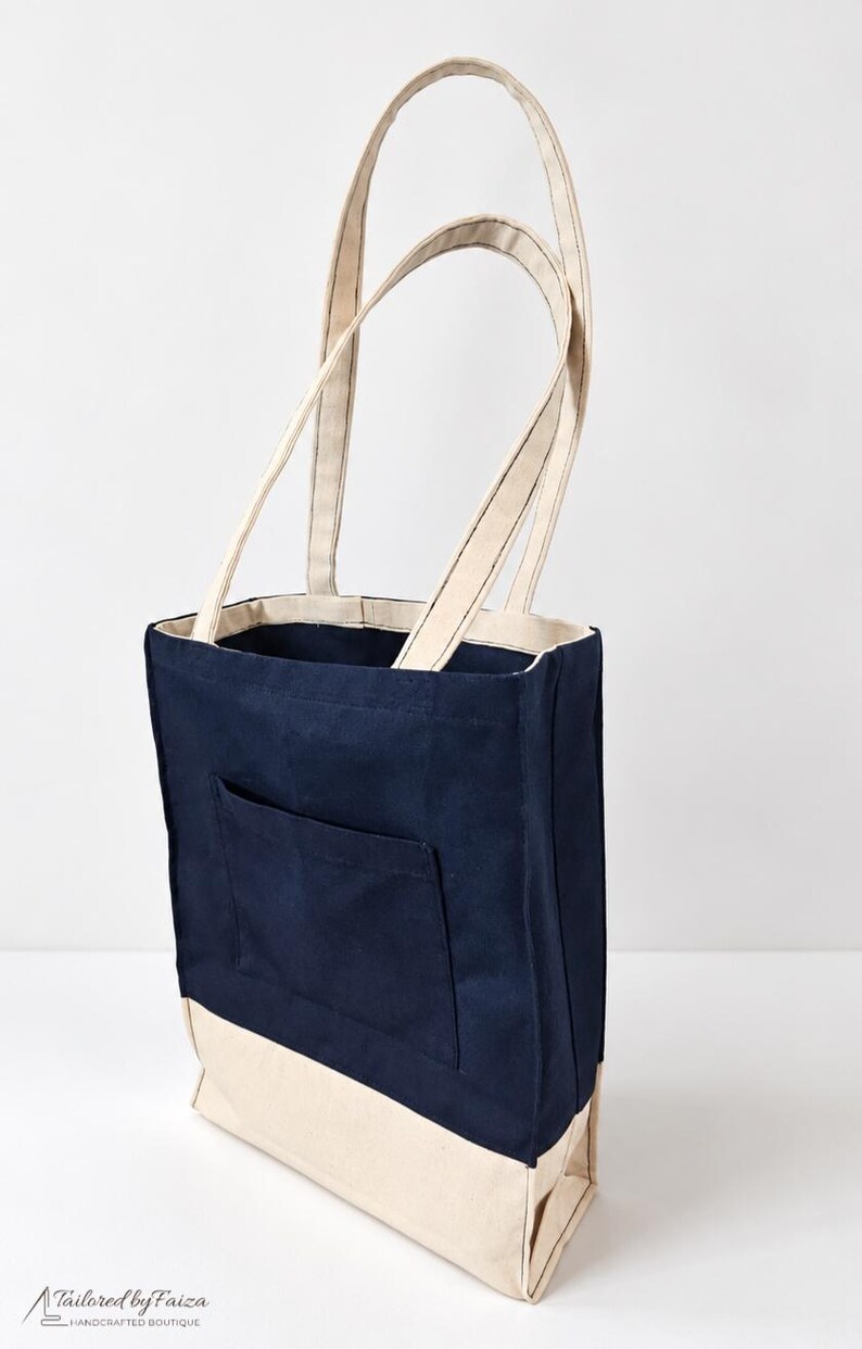 Puede incluir: Un bolso tote de lona en azul marino y crema con dos asas largas para el hombro, un bolsillo exterior rectangular en la parte delantera y una base s&oacute;lida de color crema. En la esquina inferior izquierda aparece el texto Tailored by Faiza Handcrafted Boutique.