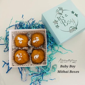May include: A light blue box with the text "It's a Boy" cut out on the lid, next to a white box containing four round, orange-colored sweets. The sweets are decorated with silver leaf. The text "Tailored By Faiza" and "Baby Boy Mithai Boxes" is also visible.