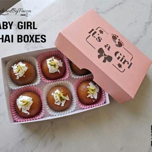 May include: A rectangular pink box with the text It's a Girl and baby footprint graphics, containing six round brown sweets in individual paper liners. Each sweet is topped with white flakes and green slivers. Text reads BABY GIRL MITHAI BOXES.