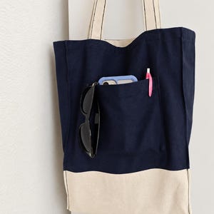 Puede incluir: Un bolso de tela azul marino con un panel inferior de color crema y dos correas largas para el hombro. La parte delantera tiene un bolsillo grande que contiene un tel&eacute;fono inteligente azul claro, unas gafas de sol negras y un bol&iacute;grafo rosa. El texto Tailored by Faiza Handcrafted Boutique es visible en la esquina inferior izquierda.