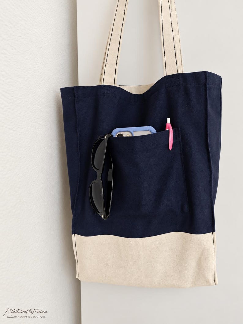 Puede incluir: Un bolso de tela azul marino con un panel inferior de color crema y dos correas largas para el hombro. La parte delantera tiene un bolsillo grande que contiene un tel&eacute;fono inteligente azul claro, unas gafas de sol negras y un bol&iacute;grafo rosa. El texto Tailored by Faiza Handcrafted Boutique es visible en la esquina inferior izquierda.