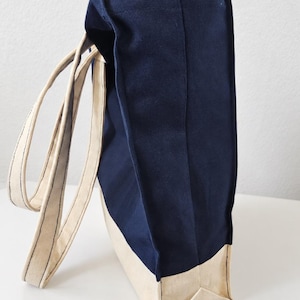 Puede incluir: Un bolso tote de lona en azul marino y crema con dos asas largas de color crema. El bolso presenta una base rectangular y un dise&ntilde;o de paneles laterales verticales. El logotipo Tailored by Faiza Handcrafted Boutique est&aacute; impreso en la esquina inferior izquierda.