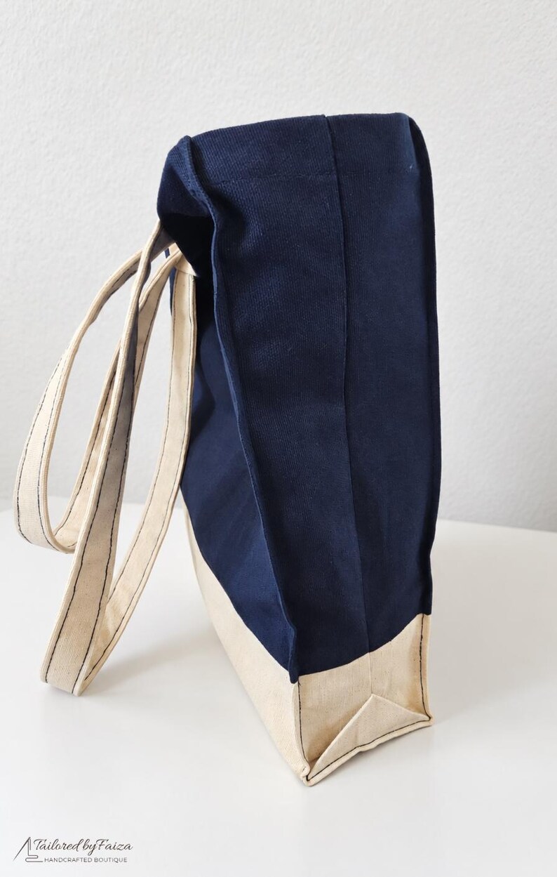 Puede incluir: Un bolso tote de lona en azul marino y crema con dos asas largas de color crema. El bolso presenta una base rectangular y un dise&ntilde;o de paneles laterales verticales. El logotipo Tailored by Faiza Handcrafted Boutique est&aacute; impreso en la esquina inferior izquierda.