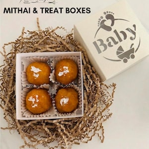 May include: A white box with the word "Baby" and a stroller graphic, next to a box of four orange mithai treats. The treats are in a white box, nestled in brown paper. The text "Mithai & Treat Boxes" is at the top.