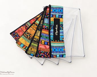 Puede incluir: Un conjunto de cinco estuches de vinilo transparente dispuestos en abanico. Cada estuche tiene una cremallera negra y un borde de tela con un patrón geométrico colorido. Un estuche lleva una etiqueta negra con el nombre Sophia en blanco.