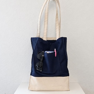 Puede incluir: Un bolso tote azul marino con una base de color crema y asas a juego. El bolsillo frontal contiene unas gafas de sol negras, un tel&eacute;fono inteligente y un bol&iacute;grafo rosa. El texto Tailored by Faiza est&aacute; impreso en la parte inferior.