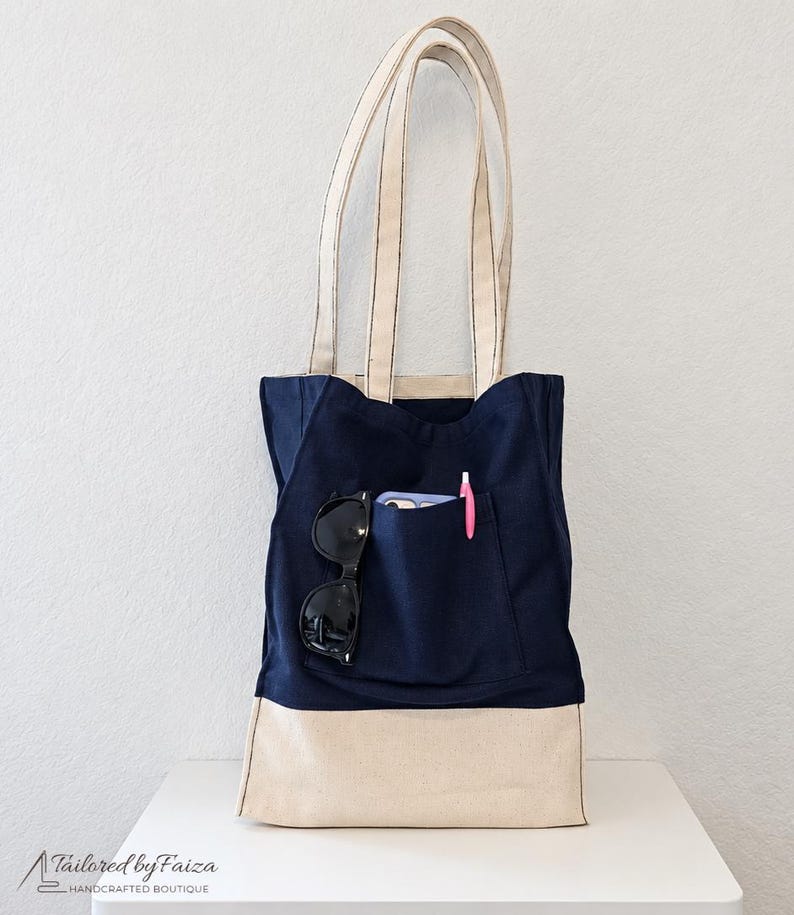 Puede incluir: Un bolso tote azul marino con una base de color crema y asas a juego. El bolsillo frontal contiene unas gafas de sol negras, un tel&eacute;fono inteligente y un bol&iacute;grafo rosa. El texto Tailored by Faiza est&aacute; impreso en la parte inferior.