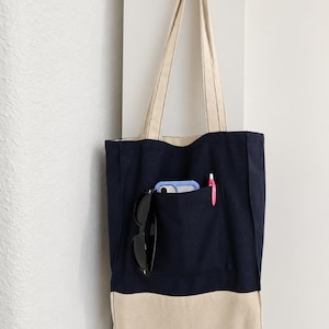 Puede incluir: Un bolso tote de lona azul marino con un panel inferior beige y dos correas largas para el hombro. La parte delantera cuenta con un bolsillo grande que contiene un smartphone azul claro, gafas de sol negras y un bol&iacute;grafo rosa. El texto Tailored by Faiza es visible.