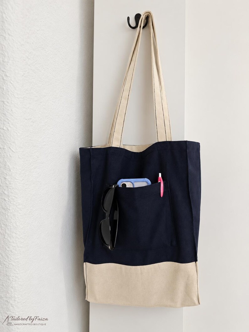Puede incluir: Un bolso tote de lona azul marino con un panel inferior beige y dos correas largas para el hombro. La parte delantera cuenta con un bolsillo grande que contiene un smartphone azul claro, gafas de sol negras y un bol&iacute;grafo rosa. El texto Tailored by Faiza es visible.