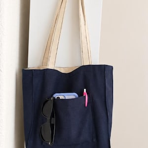 Puede incluir: Un bolso tote de lona en azul marino y beige con correas largas para el hombro. La parte frontal tiene un bolsillo grande que contiene un smartphone azul claro, gafas de sol negras y un bol&iacute;grafo rosa. La base es beige y la parte superior azul.