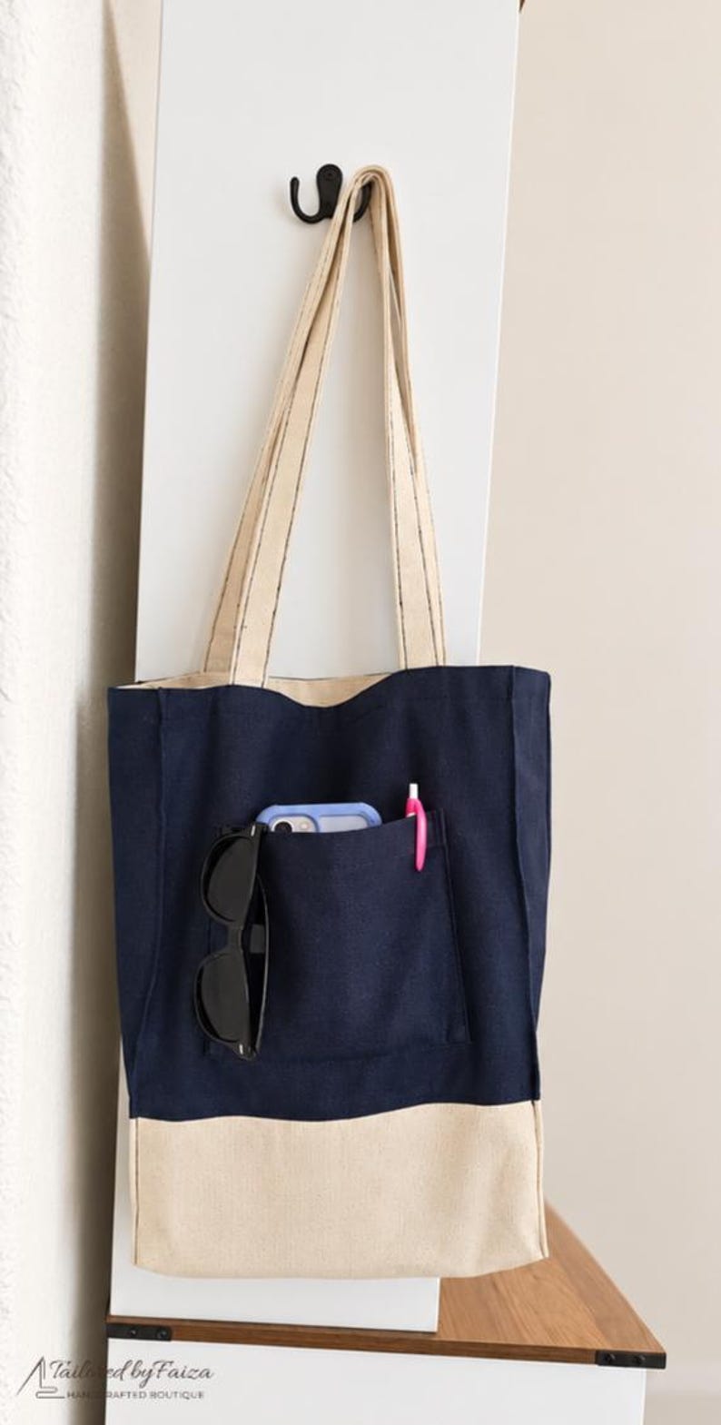 Puede incluir: Un bolso tote de lona en azul marino y beige con correas largas para el hombro. La parte frontal tiene un bolsillo grande que contiene un smartphone azul claro, gafas de sol negras y un bol&iacute;grafo rosa. La base es beige y la parte superior azul.