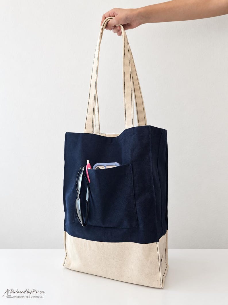 Puede incluir: Un bolso tote de lona en azul marino y crema con dos asas largas para el hombro. La parte frontal tiene un bolsillo exterior grande que contiene un tel&eacute;fono inteligente, un bol&iacute;grafo rosa y gafas de montura negra. La base es de color crema.