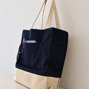 Puede incluir: Un bolso tote de lona azul marino y crema con dos asas largas para el hombro. El bolso cuenta con un bolsillo exterior grande que contiene un bol&iacute;grafo rosa y un smartphone morado claro. Unas gafas de sol negras cuelgan del borde. Tailored by Faiza.