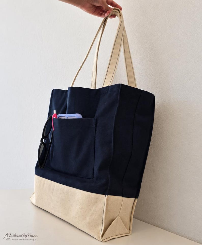 Puede incluir: Un bolso tote de lona azul marino y crema con dos asas largas para el hombro. El bolso cuenta con un bolsillo exterior grande que contiene un bol&iacute;grafo rosa y un smartphone morado claro. Unas gafas de sol negras cuelgan del borde. Tailored by Faiza.