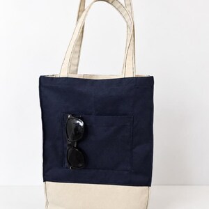 Puede incluir: Un bolso tote de lona azul marino con una base y asas de color crema a juego. El bolso cuenta con un bolsillo exterior frontal que contiene un par de gafas de sol negras. El texto Tailored by Faiza Handcrafted Boutique es visible.