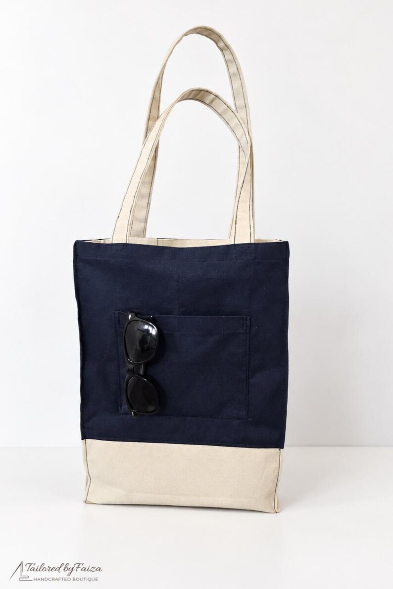 Puede incluir: Un bolso tote de lona azul marino con una base y asas de color crema a juego. El bolso cuenta con un bolsillo exterior frontal que contiene un par de gafas de sol negras. El texto Tailored by Faiza Handcrafted Boutique es visible.