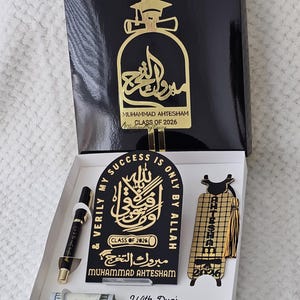 May include: A personalized deluxe Islamic graduation gift set in a black box. The set includes a pen, a bookmark, and a plaque with Arabic calligraphy and the name "Muhammad Ahtesham" and "Class of 2026". The box has a gold graduation cap design.