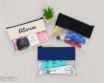 Puede incluir: Tres estuches rectangulares con cremallera, cuerpo de vinilo transparente y parte superior de tela de colores. Un estuche es crema con el nombre Olivia impreso en negro, otro tiene la parte superior negra y otro azul marino.