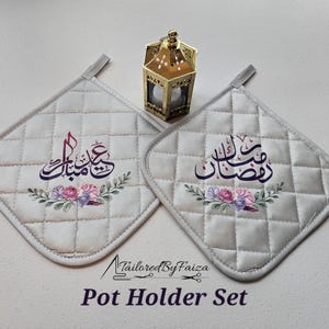 May include: A set of two white quilted pot holders with floral designs and Arabic calligraphy. The pot holders feature the words "Eid Mubarak" and "Ramadan Kareem" in purple and pink lettering.
