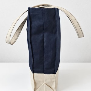 Puede incluir: Un bolso tote vertical con una secci&oacute;n superior azul marino profundo y una base inferior beige en contraste. El bolso incluye dos asas de tela beige y costuras visibles a lo largo de los lados. El texto Tailored by Faiza Handcrafted Boutique est&aacute; impreso en la esquina inferior izquierda.